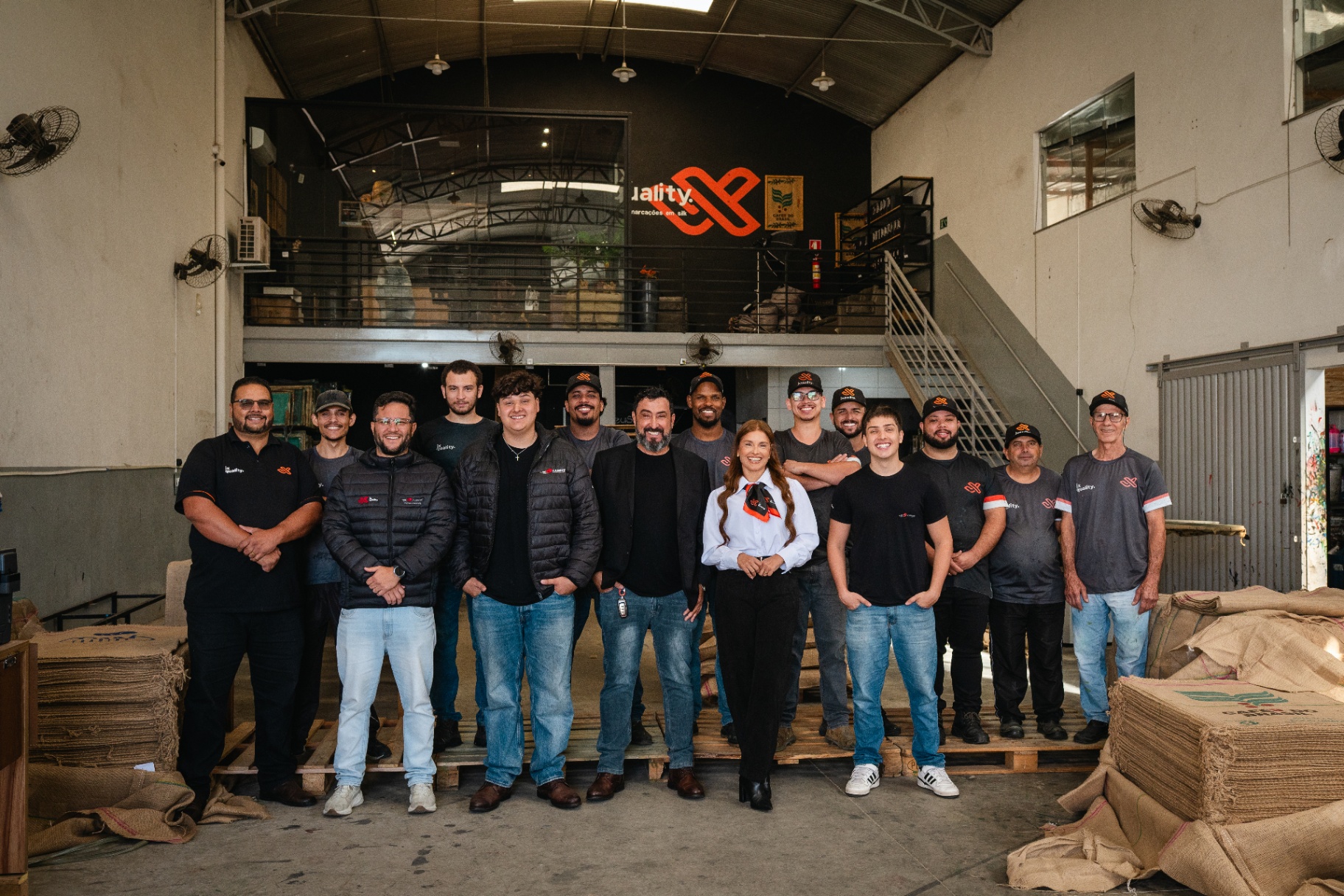 Equipe JX Quality reunida no galpão da operação em Varginha, com sacaria de juta em primeiro plano e o logo JX ao fundo